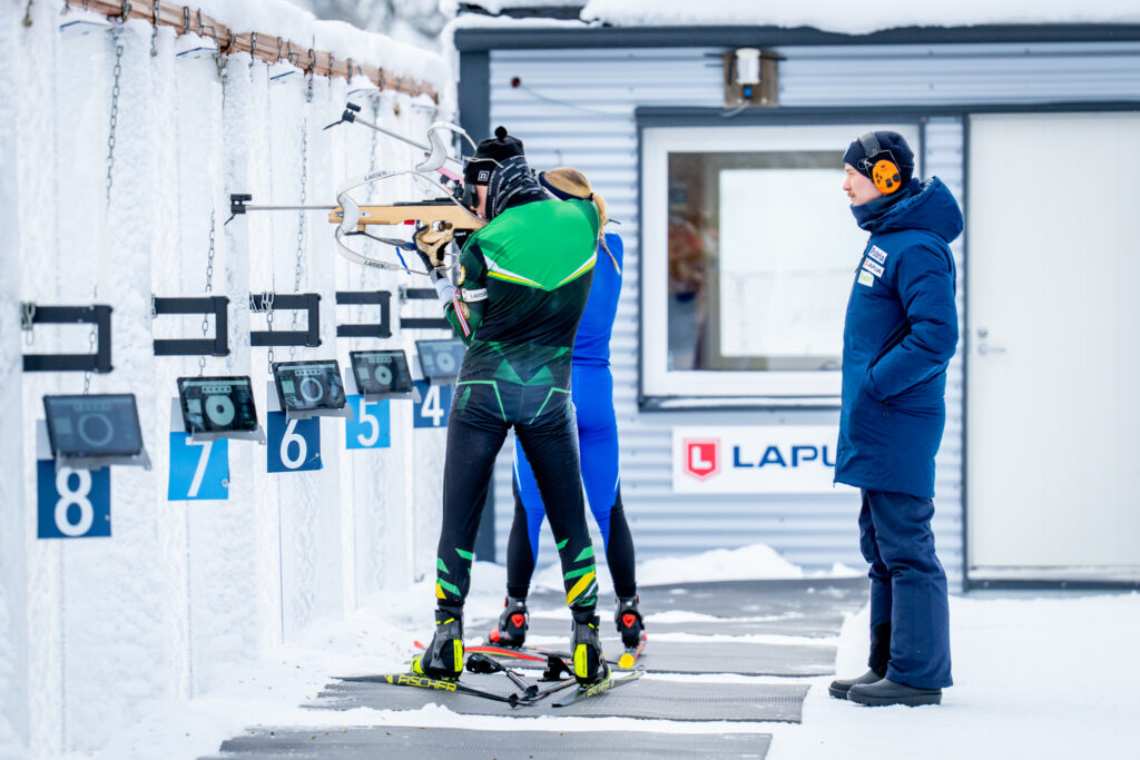 Kuvassa kaksi ampumahiihtäjää on ampumapenkalla Vuokatissa konttilaboratoriolla ja valmentaja seisoo heidän takanaan.