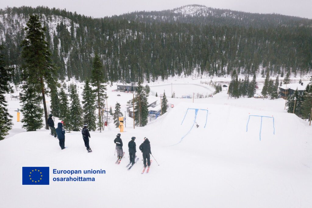 Kuva Rukan laskettelukeskuksen RUKA PARK rinteestä, jossa seisoo laskettelijoita suksien päällä rinteen yläpäässä. EU:n osarahoittama logo vasemmassa alakulmassa.