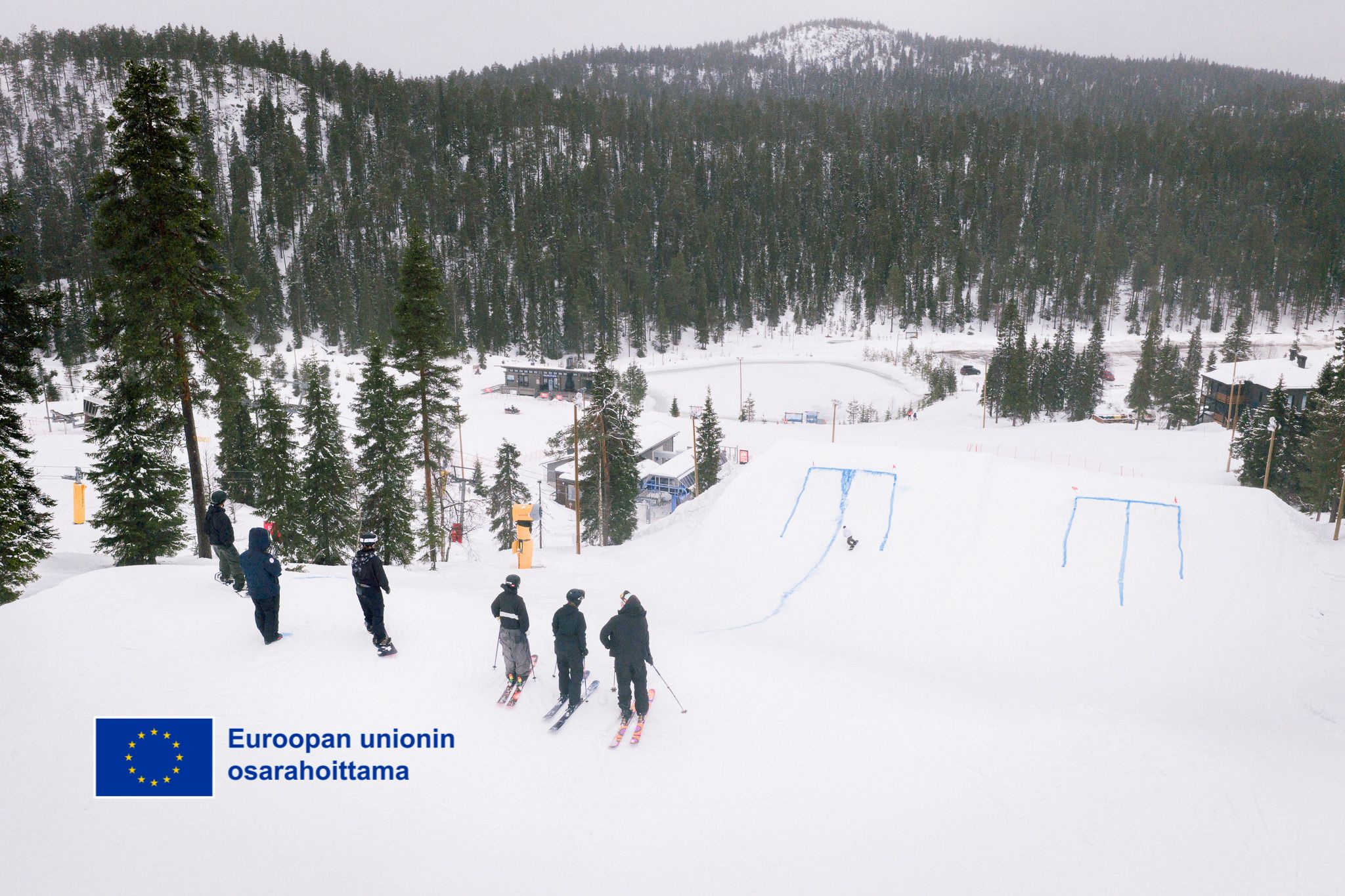 Kuva Rukan laskettelukeskuksen RUKA PARK rinteestä, jossa seisoo laskettelijoita suksien päällä rinteen yläpäässä. EU:n osarahoittama logo vasemmassa alakulmassa.