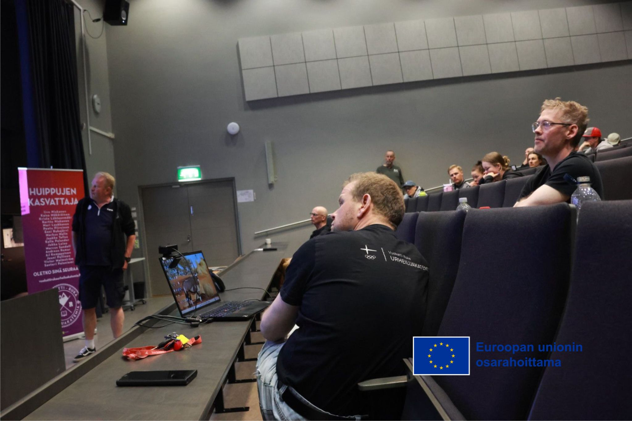 Kuvassa Vuokatti-Ruka urheiluakatemian henkilöstöä Kuusamon lukion auditoriossa istumassa. Kuvassa näkyy myös valkokangas ja reuna siihen heijastetusta materiaalista. Valkokankaan edessä seisoo tilaisuutta vetänyt Olli-Pekka Kärkkäinen.