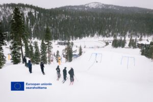 Kuva Rukan laskettelukeskuksen RUKA PARK rinteestä, jossa seisoo laskettelijoita suksien päällä rinteen yläpäässä. EU:n osarahoittama logo vasemmassa alakulmassa.