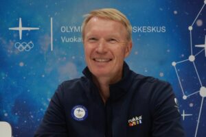 Tapio Korjus hymyilee. Taustalla on Olympiavalmennuskekus Vuokatti-Rukan seinä.